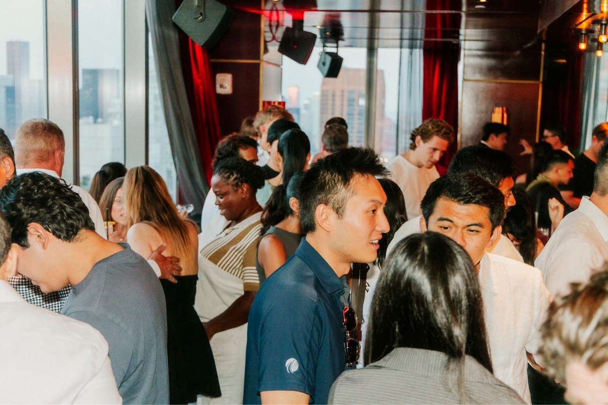 A full room of guests mingling under warm red-gold light at the NYC Tech Week Rooftop Mixer